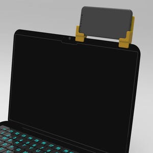 May include: A black laptop with a teal backlit keyboard and a yellow phone holder attached to the top of the screen. A grey smartphone is placed in the holder. The laptop is on a white background.