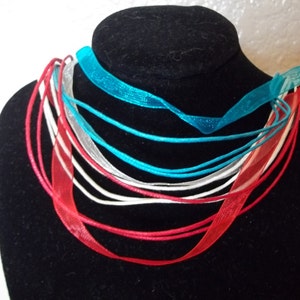 May include: A multi-strand necklace displayed on a black mannequin. The necklace features strands of red, white, silver, and turquoise ribbons and cords. The ribbons are sheer and the cords are solid. The necklace is a fashion accessory.