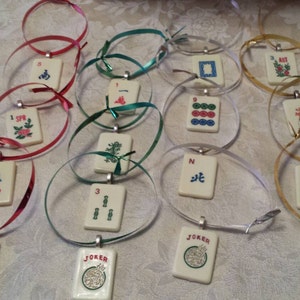 May include: A collection of ivory-colored Mahjong tile pendants with various symbols and numbers, strung on colorful ribbons. The ribbons are in shades of red, green, silver, and gold. Each pendant has a silver-colored bail. Some pendants have the word "JOKER".