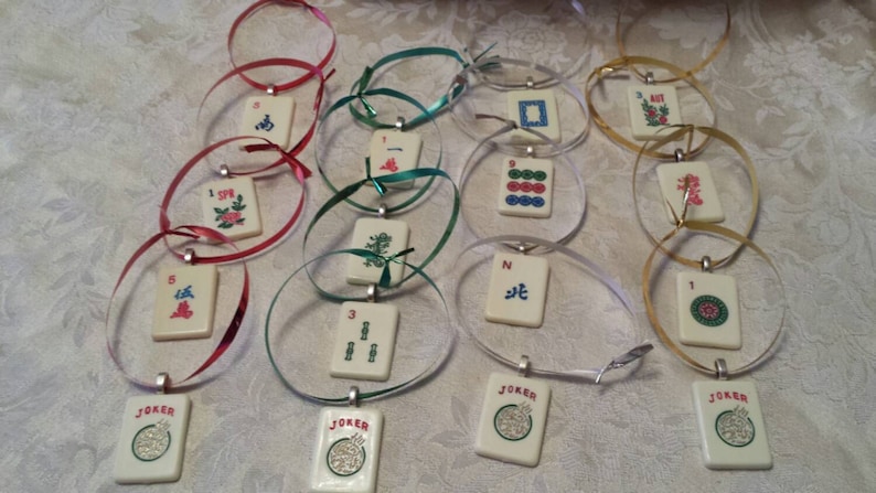 May include: A collection of ivory-colored Mahjong tile pendants with various symbols and numbers, strung on colorful ribbons. The ribbons are in shades of red, green, silver, and gold. Each pendant has a silver-colored bail. Some pendants have the word "JOKER".
