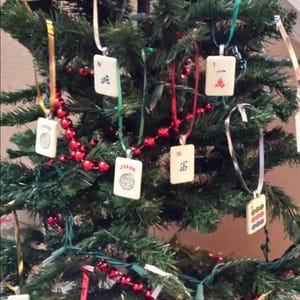 May include: A collection of off-white rectangular Mahjong tiles used as hanging ornaments on a green artificial Christmas tree. Each tile features unique black, red, or blue ink markings, including the word JOKER, Chinese characters, and numbers.