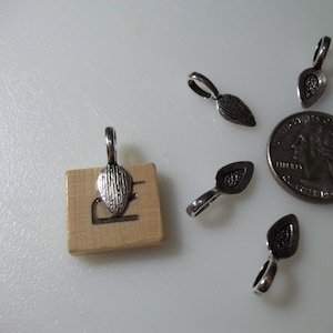 May include: A close-up of silver-toned metal pendant bails, with a textured, teardrop-shaped design. The bails are displayed on a wooden surface, with a US quarter for scale.