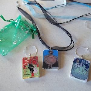 May include: Three rectangular pendants with images from The Wizard of Oz, each attached to a silver hoop and black ribbon. A green mesh gift bag with silver accents is also present, along with additional ribbons.
