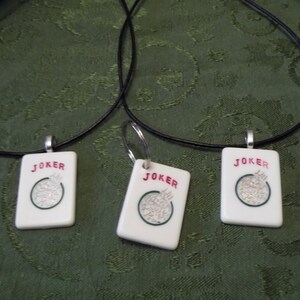 May include: Three black cord necklaces with white ceramic pendants shaped like playing cards. Each pendant has the word "JOKER" printed in red and a green circle with a white design in the center.