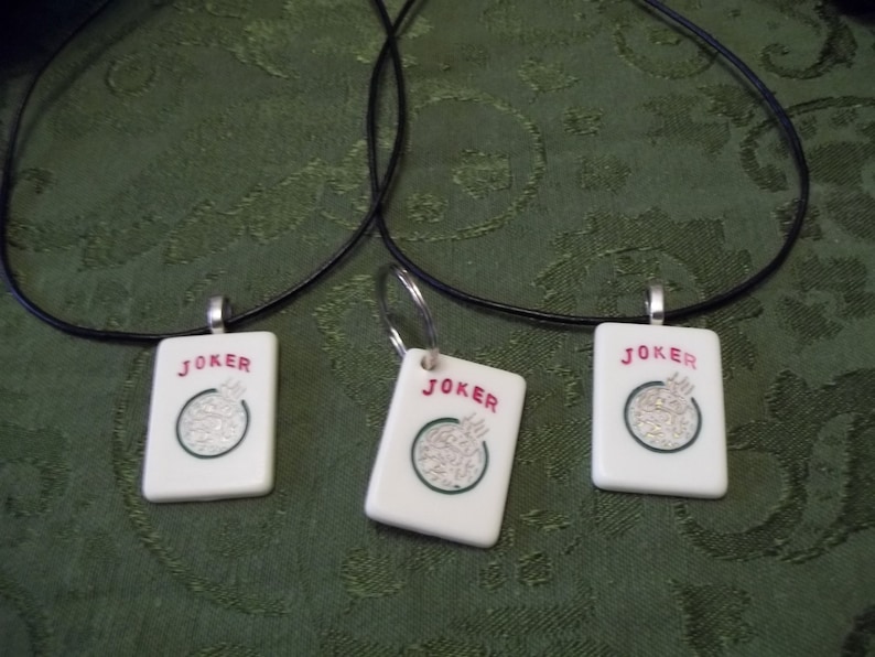 May include: Three black cord necklaces with white ceramic pendants shaped like playing cards. Each pendant has the word "JOKER" printed in red and a green circle with a white design in the center.