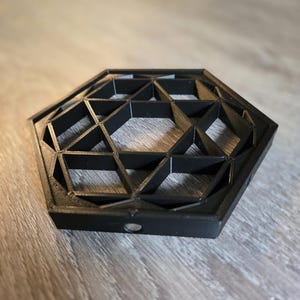 May include: A black hexagonal coaster featuring an intricate, raised geometric lattice pattern composed of interconnected triangles and hexagons. The object has a solid outer frame and a small circular opening visible on the front side edge.