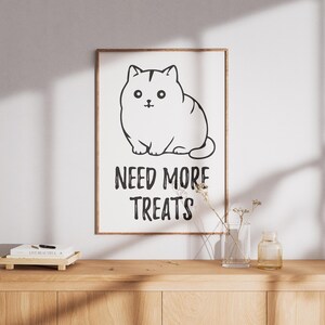 Könnte beinhalten: Ein rechteckiger Wandkunstdruck in einem dünnen Holzrahmen mit einer schwarz-weißen Strichzeichnung einer runden Katze. Unter der Katzenillustration steht der Text NEED MORE TREATS in fetten, schwarzen Großbuchstaben auf weißem Hintergrund.