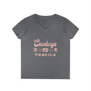May include: A heather gray V-neck t-shirt with the text "Cowboys Tequila" in a pink script font. The design includes pink decorative elements and the words "Legend Since 1975".