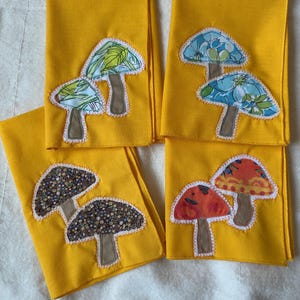 Puede incluir: Cuatro servilletas de tela de color amarillo brillante, cada una con dos diseños de setas aplicados. Las setas tienen tallos marrones y sombreros estampados en varios colores, incluyendo verde, azul, naranja y estampados florales oscuros.