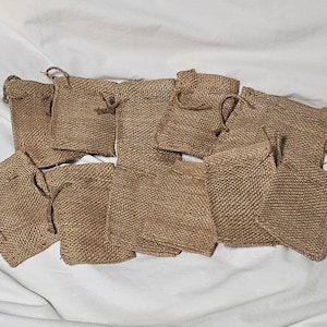 May include: A collection of ten small, rectangular drawstring pouches crafted from a coarse, tan-colored burlap fabric. Each pouch features a woven texture and a matching jute string closure at the top, arranged in a loose cluster.
