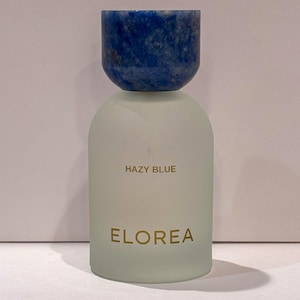 Peut inclure: Un flacon de parfum cylindrique avec un corps en verre d&eacute;poli et un bouchon arrondi en pierre bleu fonc&eacute;. Le texte HAZY BLUE est imprim&eacute; en dor&eacute; au-dessus du nom de marque ELOREA, &eacute;galement imprim&eacute; en dor&eacute; sur le devant du flacon en verre.