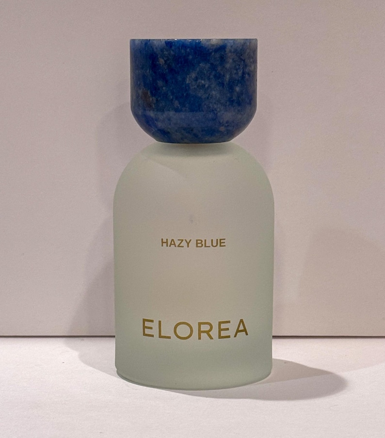 Peut inclure: Un flacon de parfum cylindrique avec un corps en verre d&eacute;poli et un bouchon arrondi en pierre bleu fonc&eacute;. Le texte HAZY BLUE est imprim&eacute; en dor&eacute; au-dessus du nom de marque ELOREA, &eacute;galement imprim&eacute; en dor&eacute; sur le devant du flacon en verre.