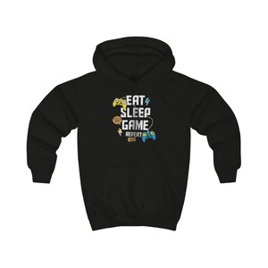 Peut inclure: Un sweat-shirt à capuche noir avec le texte blanc EAT SLEEP GAME REPEAT. Le motif comprend une manette de jeu vidéo jaune, une manette bleue, ainsi que de petites icônes graphiques orange et jaunes incluant des éclairs et des bulles.