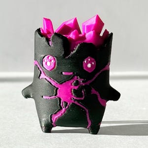 May include: A small, three-dimensional printed figurine featuring a matte black cylindrical body with jagged edges at the top. Bright magenta crystalline structures emerge from the center, while magenta vein-like patterns and two eyes decorate the front.