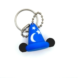 Op de afbeelding: Een blauwe tovenaarsmuts sleutelhanger met een wit patroon van een maansikkel en sterren op de kegel. De muts heeft twee zwarte ronde oren aan de zijkanten en is bevestigd aan een zilverkleurige metalen ring en een kleine zilveren kogelketting.