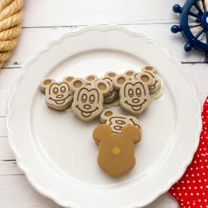 May include: A collection of light brown, Mickey Mouse-shaped treats arranged on a white scalloped plate. Several pieces feature a detailed embossed face of the character, while one piece in the foreground is plain with a small yellow centre dot.
