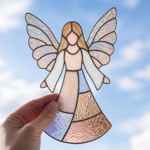 May include: A stained glass angel ornament featuring iridescent wings and a multi-colored dress composed of textured glass panels in shades of pale pink, soft blue, and cream, all held together by dark metal solder lines in a classic silhouette.