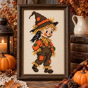 May include: A framed cross-stitch embroidery featuring a scarecrow character wearing a dark brown hat with an orange band, a bright orange shirt, and dark brown overalls with orange patches, set against a light cream-colored fabric background.