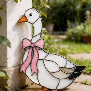 May include: A stained glass goose figure featuring white glass panels for the body, an orange beak, and a black eye. The goose is adorned with a pink glass bow around its neck and has black glass accents on the tail feathers, all held together by dark metal solder lines.