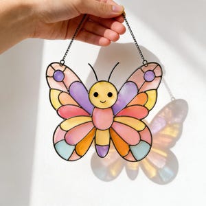 Peut inclure: Un attrape-soleil papillon en vitrail composé d'une tête jaune avec des yeux noirs et un sourire, entourée d'ailes formées de segments de verre translucide aux tons pastel rose, violet, orange, jaune et bleu clair, suspendu par une chaîne.