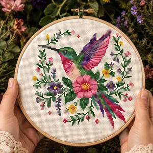 May include: A circular wooden embroidery hoop containing a cross-stitch pattern on white fabric. The design features a central hummingbird with green, pink, and purple plumage, surrounded by a wreath of small colorful flowers and green leaves.