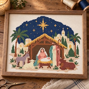 May include: A framed cross-stitch embroidery depicting a nativity scene with a central stable, a bright yellow star in a dark blue night sky, palm trees, and figures including a donkey, a sheep, and a cow on a light fabric background.