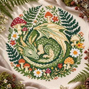 Op de afbeelding: Een kruissteekborduurwerk op crèmekleurige stof met een opgerolde groene draak omringd door een cirkelvormige schikking van donkergroene varenbladeren, witte madeliefjes en diverse rode, oranje en crèmekleurige paddenstoelen in een patroon.