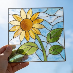 May include: A square stained glass panel featuring a central yellow sunflower with a textured orange center, green leaves on a stem, and a light blue and white swirled background, all held together by thin dark metal lead lines in a frame.