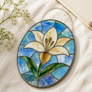 Könnte beinhalten: Ein ovaler Buntglas-Sonnenfänger mit einer zentralen weißen und gelben Lilienblüte und grünen Blättern vor einem Hintergrund aus wirbelnden blauen und weißen Glasstücken, eingefasst in dunkles Metall mit einer silberfarbenen Aufhängungskette.