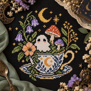 Könnte beinhalten: Eine Kreuzstichstickerei auf schwarzem Stoff mit einem weißen Geist, der in einer weißen Teetasse mit blauen Blumenmustern und einem goldenen Halbmond sitzt. Das Design umfasst bunte Pilze, violette Glockenblumen und einen gelben Mond.