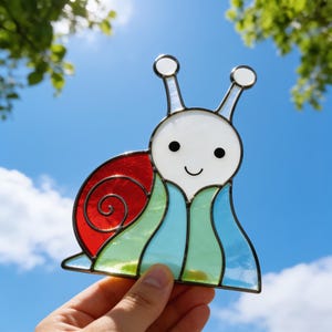 Puede incluir: Un atrapasoles de vidriera en forma de caracol con cabeza blanca, ojos negros y boca sonriente, una concha roja en espiral y un cuerpo compuesto por segmentos de vidrio verde claro y azul claro unidos por líneas de soldadura oscuras.