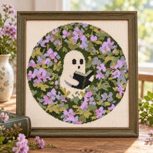May include: A framed cross-stitch embroidery featuring a white ghost reading a black book, centered within a circular pattern of green leaves and purple flowers. The artwork is enclosed in a weathered green wooden frame with a square profile.
