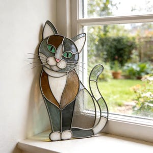May include: A stained glass cat suncatcher featuring a calico pattern of brown, gray, and white glass segments. The cat has bright green eyes, a small pink nose, and thin metal wire whiskers, all held together by dark metal solder lines.