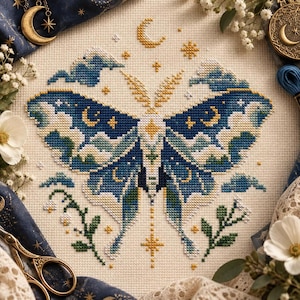 Può includere: Un ricamo a punto croce su tessuto color crema raffigurante una grande falena con ali decorate in tonalità blu scuro, verde acqua e crema. Il disegno include motivi celesti come lune e stelle, con piccoli rametti verdi alla base.