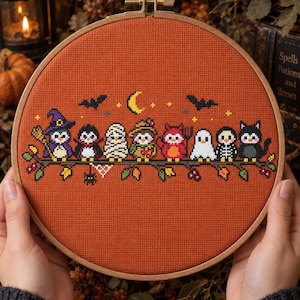 May include: A round wooden embroidery hoop containing a cross-stitch design on burnt orange fabric. The pattern features eight small characters dressed in Halloween costumes, including a witch, mummy, ghost, and black cat, perched on a branch.