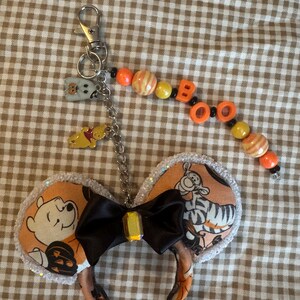 May include: A keychain featuring a miniature pair of mouse ears with orange fabric printed with cartoon characters, a black satin bow with a yellow gem, and a silver metal clasp. A beaded strand spells BOO with orange and yellow beads.