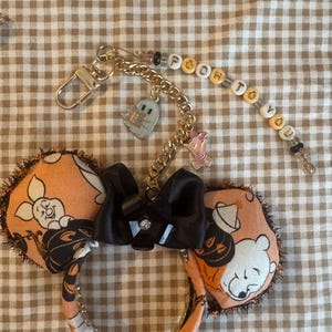 May include: A headband featuring orange fabric with a Winnie the Pooh and Piglet pumpkin print, a black satin bow, and a gold-tone chain attachment. The chain includes a ghost charm, a Piglet charm, and beads spelling out POOH TO YOU.