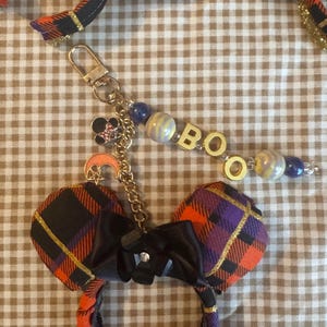 May include: A keychain featuring a miniature pair of mouse ears in an orange, purple, and black plaid fabric with a black satin bow. It includes a gold-tone metal clasp, a crescent moon charm, a mouse head charm, and beads spelling the word BOO.