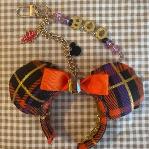 May include: A headband featuring two round ears covered in a black, orange, and purple plaid fabric with gold glitter accents. A bright orange ribbon bow sits in the center. A gold chain attachment features a red leaf charm, a black mouse head icon, and a beaded strand with gold letter beads spelling BOO.