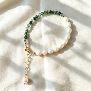 May include: A bracelet featuring a strand of small, round, dark green moss agate beads and larger, white, oval-shaped freshwater pearls. The piece includes a gold-toned metal lobster clasp and an extender chain with heart-shaped links and a pearl.