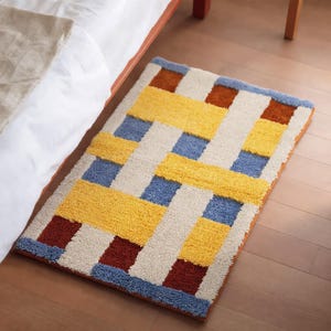 May include: A rectangular area rug featuring a woven-look pattern with a textured pile. The design consists of interlocking blocks in shades of mustard yellow, cream, deep rust brown, and muted blue, arranged in a repeating geometric grid layout.
