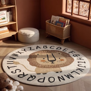 May include: A round, cream-colored area rug featuring a central illustration of a brown lion with a dark brown mane. The alphabet from A to Z is printed in a black, sans-serif font in a circular pattern around the perimeter of the rug.