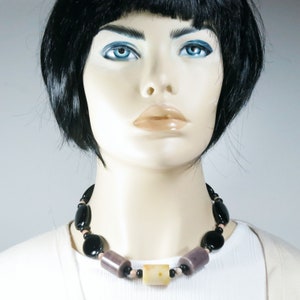 May include: A necklace featuring a mix of black, purple, and cream-colored beads. The necklace has a variety of shapes, including round and cylindrical beads. The necklace is displayed on a mannequin.