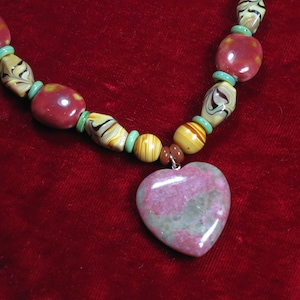 May include: A heart-shaped pendant necklace featuring a pink and green stone. The necklace is crafted with brown, yellow, and white beads with green accents.