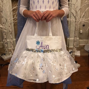 May include: White canvas tote bag with a silver sequin trim and a snowflake design. The bag is personalized with the name "Ava" in purple embroidery.