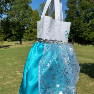 Puede incluir: Un bolso tote blanco y turquesa que se asemeja a un vestido de princesa. La parte superior es blanca con una inicial bordada en turquesa. La falda es de sat&eacute;n turquesa con una superposici&oacute;n transparente con copos de nieve plateados. Lentejuelas plateadas adornan la cintura.
