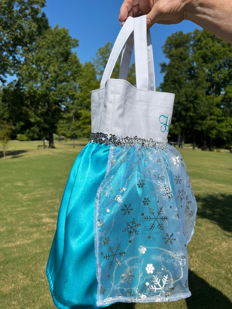 Puede incluir: Un bolso tote blanco y turquesa que se asemeja a un vestido de princesa. La parte superior es blanca con una inicial bordada en turquesa. La falda es de sat&eacute;n turquesa con una superposici&oacute;n transparente con copos de nieve plateados. Lentejuelas plateadas adornan la cintura.