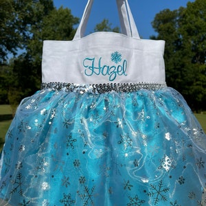 Puede incluir: Un bolso tote blanco con el nombre "Hazel" bordado en azul y un dise&ntilde;o de copo de nieve. El bolso presenta una falda tut&uacute; azul con detalles de copos de nieve plateados y un ribete de lentejuelas plateadas. El bolso se sostiene contra un fondo de &aacute;rboles verdes y cielo azul.