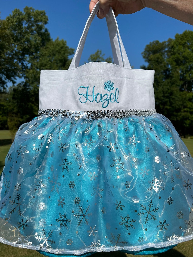 Puede incluir: Un bolso tote blanco con el nombre "Hazel" bordado en azul y un dise&ntilde;o de copo de nieve. El bolso presenta una falda tut&uacute; azul con detalles de copos de nieve plateados y un ribete de lentejuelas plateadas. El bolso se sostiene contra un fondo de &aacute;rboles verdes y cielo azul.