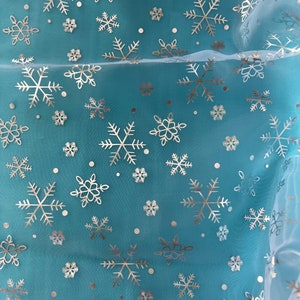 Puede incluir: Una tela transparente de color turquesa con un patr&oacute;n repetido de copos de nieve plateados y peque&ntilde;os puntos. Los copos de nieve var&iacute;an en tama&ntilde;o y dise&ntilde;o, creando un aspecto invernal y festivo. La tela es ligeramente transl&uacute;cida.